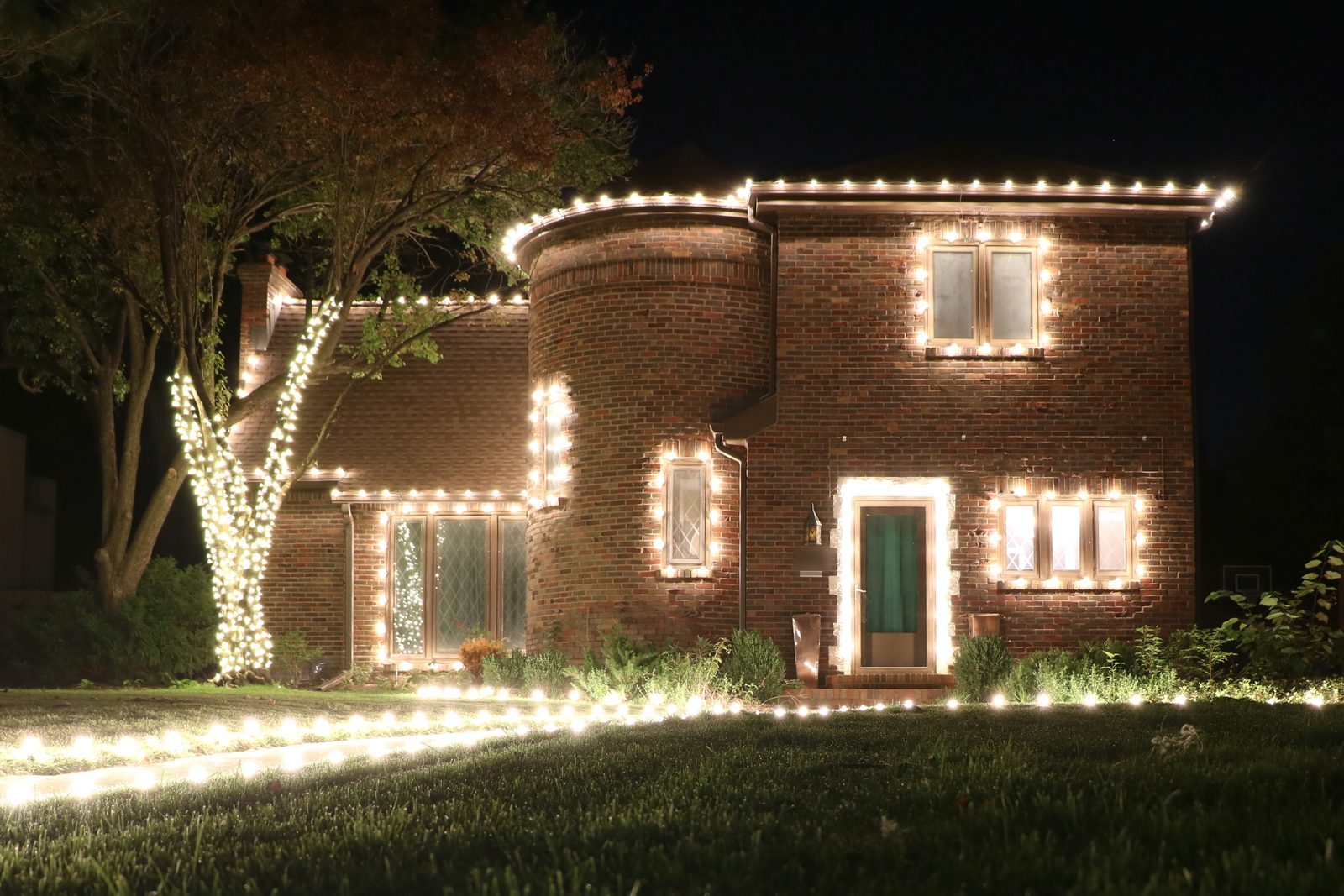 Brick home in Wichita lit with warm white roofline, window and door outlines, a wrapped tree trunk and pathway lights